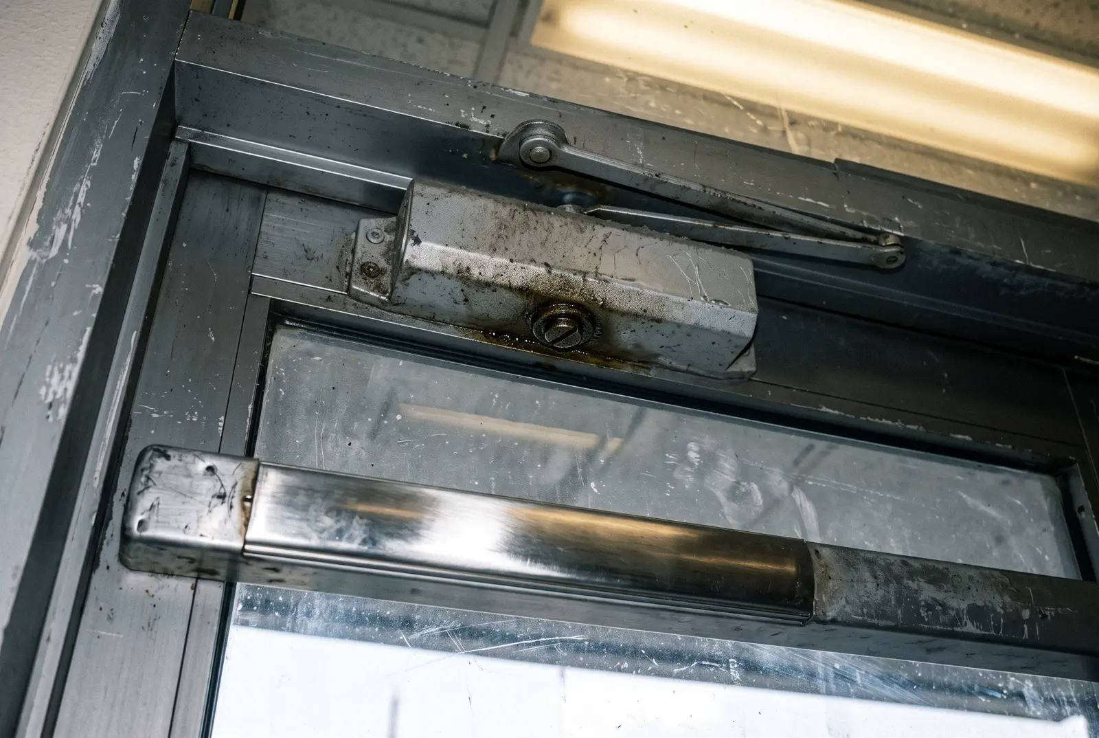 A commercial aluminium door with an exit device and a dusty overhead door closer